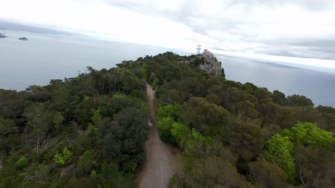 FPV drone flight up lush green mountain towards radio mast and buildings on summit overlooking Ligurian Sea near Noli, Italy. Aerial