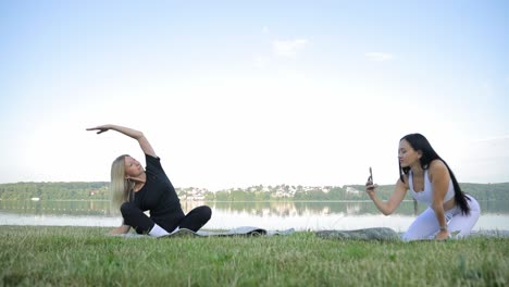 two beautiful women practicing yoga on the bank of a beautiful pond two beautiful women practicing yoga on the bank of a beautiful pond two women on gymnastic mats the girl takes pictures on the phone