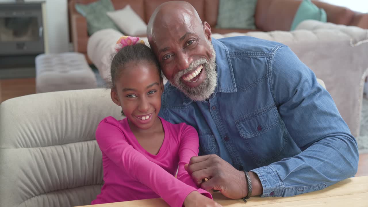 African American granddad pointing at camera, girl laughing, leaning in and bonding at home table