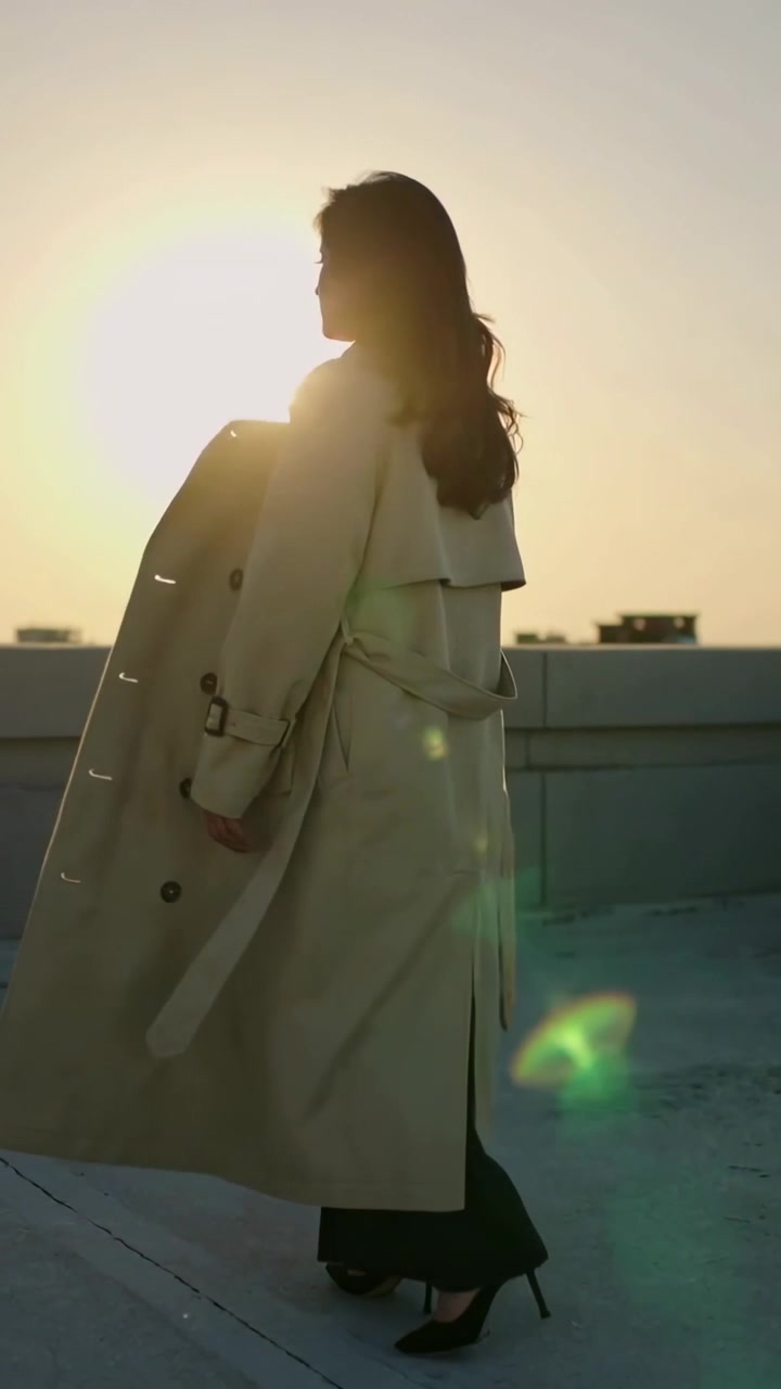 Video verticale: Donna con trench e tacchi che cammina sul parapetto del tetto mentre il sole tramonta, con bagliori.