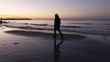 Silhouette-Eines-Mannes,-Der-Bei-Sonnenaufgang-Am-Strand-Spaziert,-Er-Hält-An,-Macht-Fotos-Und-Geht-Weiter,-Während-Ruhige-Wellen-An-Land-Kommen