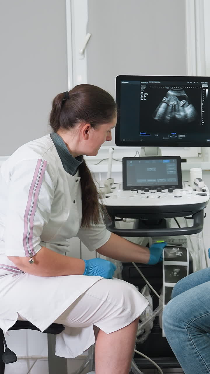 female technician performing ultrasound scan in bright clinic room, patient seated wearing jeans, sonogram fetal profile visible on monitor, technician in uniform and blue gloves operates transducer