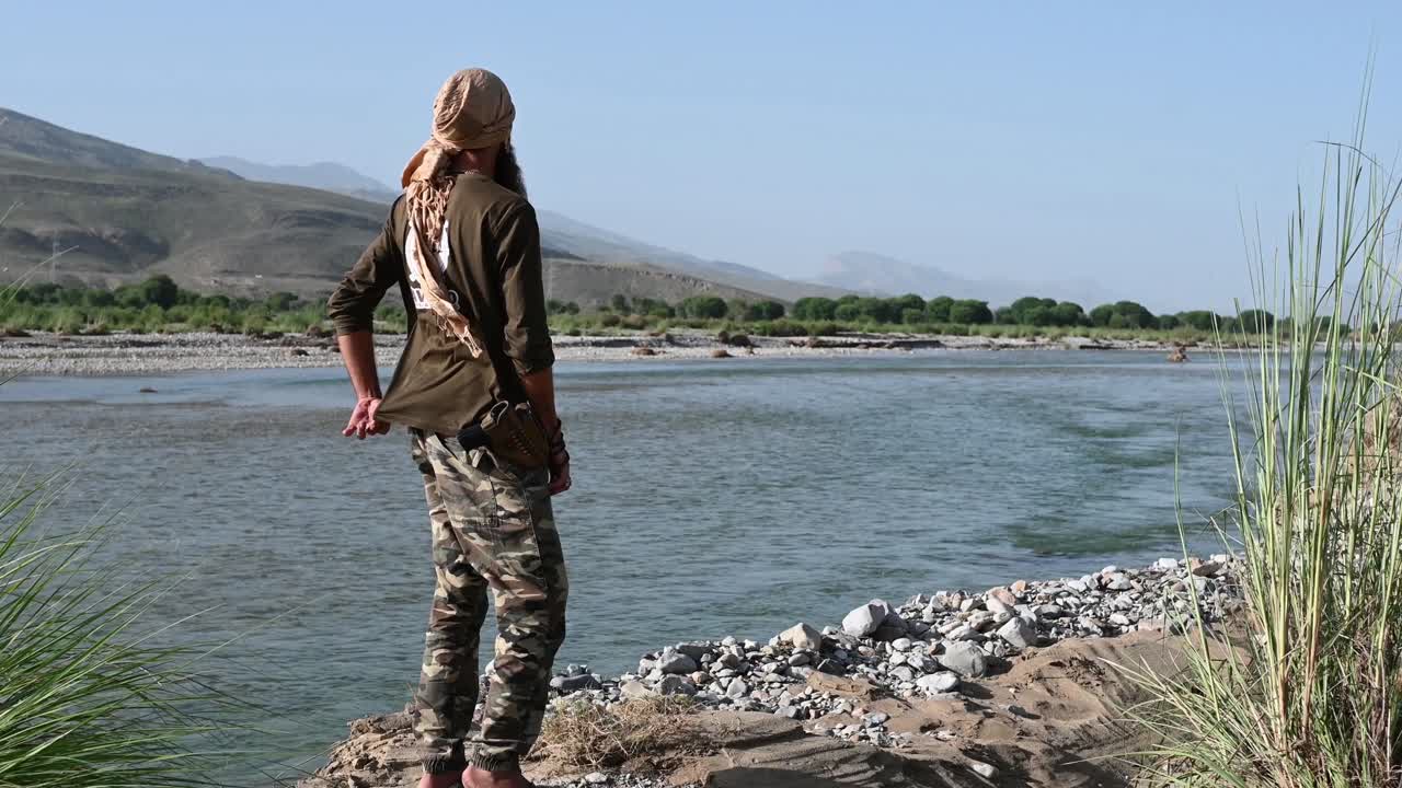 A man is standing at the edge of the river looking at the distance mountain located on the other side of the river on sunny day in Balochistan, Pakistan