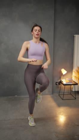 Vertical-video-of-a-confident-brunette-girl-in-a-lilac-top-and-purple-sweatpants-doing-sports-and-running-on-the-spot-at-home-in-a-modern-apartment