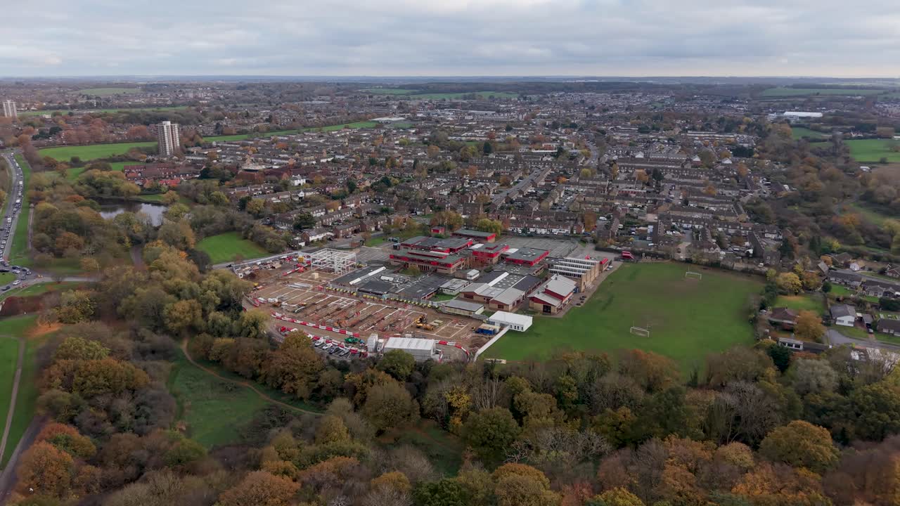 Drone flies over autumn woods towards St Mark’s Catholic School, Harlow, Essex. Ongoing construction shows sand, barriers, yellow plant machinery. A nearby road sees light traffic in both directions