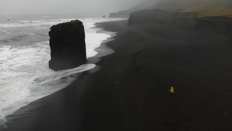 Chica-Con-Impermeable-Amarillo-Caminando-En-Una-Tormenta-A-Lo-Largo-De-Una-Playa-Negra-En-Djupavogshreppur,-Islandia