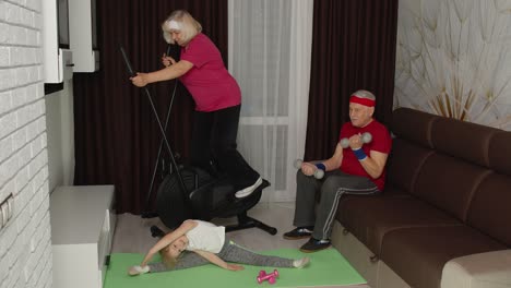 senior couple with granddaughter using orbitrek, doing weight lifting dumbbells exercises at home