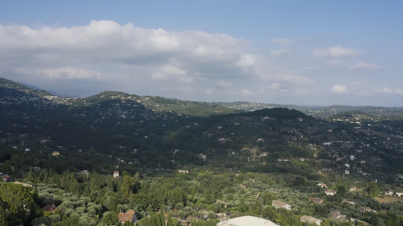 Luftfoto fra drone af bølgende bakker med spredte huse og tæt grønt over landskabet i Cagnes-sur-Mer under klart dagslys med bløde skyer på himlen.