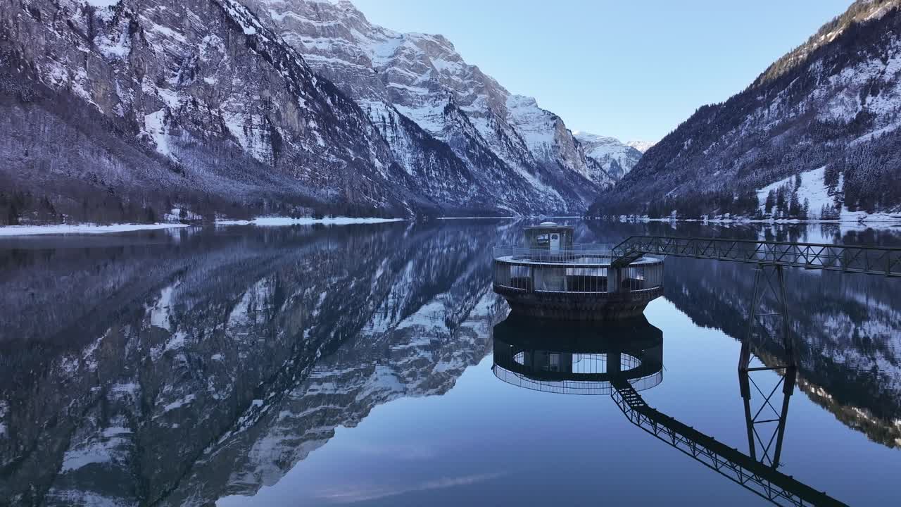 Drone perspective of the Klöntaler Wasserturm in Switzerland on a typical winter day, surrounded by snow-covered mountains and a serene frozen landscape.