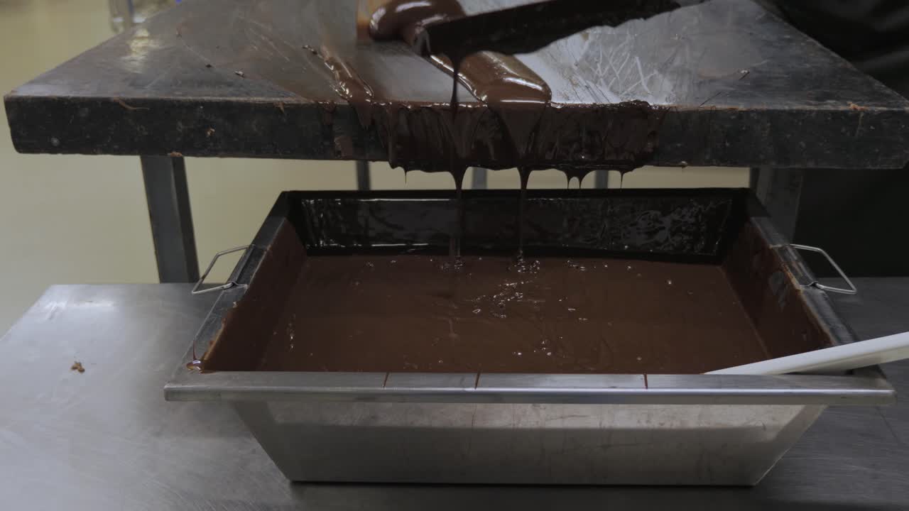 Cioccolato fondente denso e fuso che si riversa da un tavolo in un grande contenitore di metallo in un ambiente di fabbrica