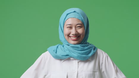팔을 고 서 있는 아시아 무슬림 여성 아보가 녹색 스크린 배경 스튜디오에서 카메라를 바라보고 미소 짓고 있다.