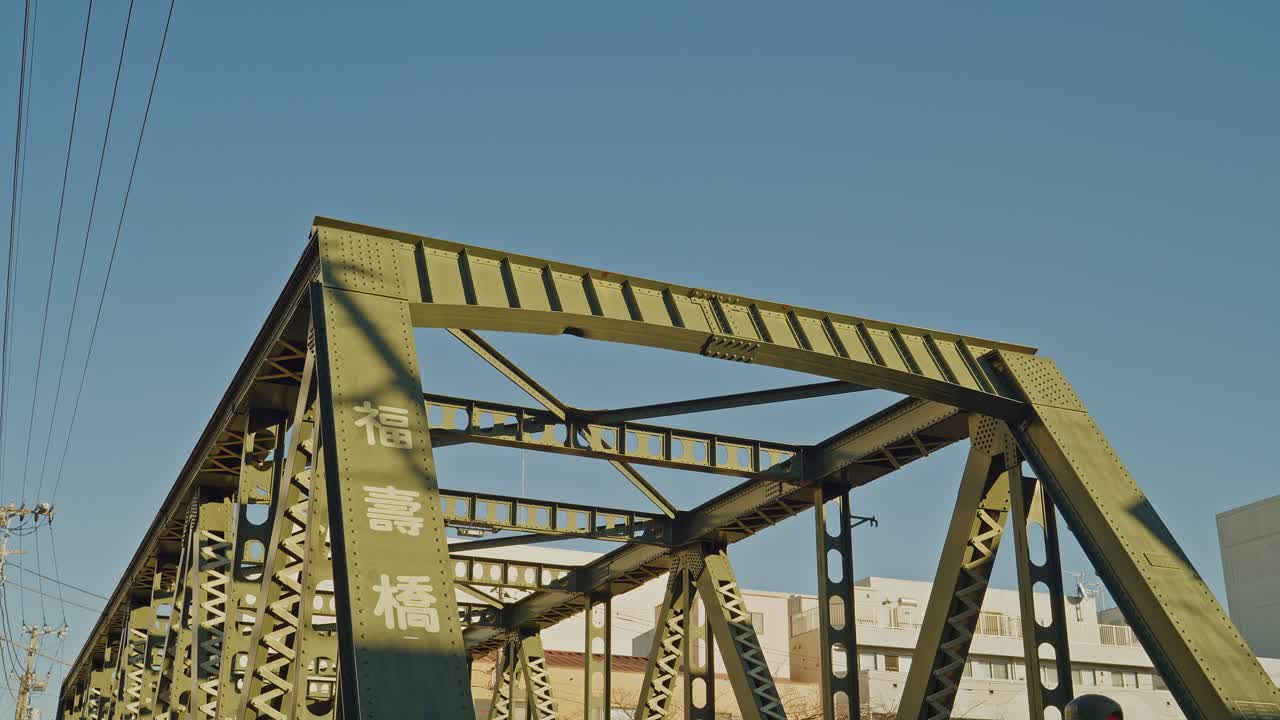 Fukuju Bridge's yellow-green steel truss fills the frame against vivid blue sky in Tokyo, with Japanese nameplate and urban buildings visible, captured as a static low-angle close-up shot