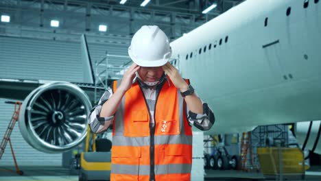 ingeniero masculino asiático con casco de seguridad que tiene dolor de cabeza mientras está de pie con aviones en el hangar