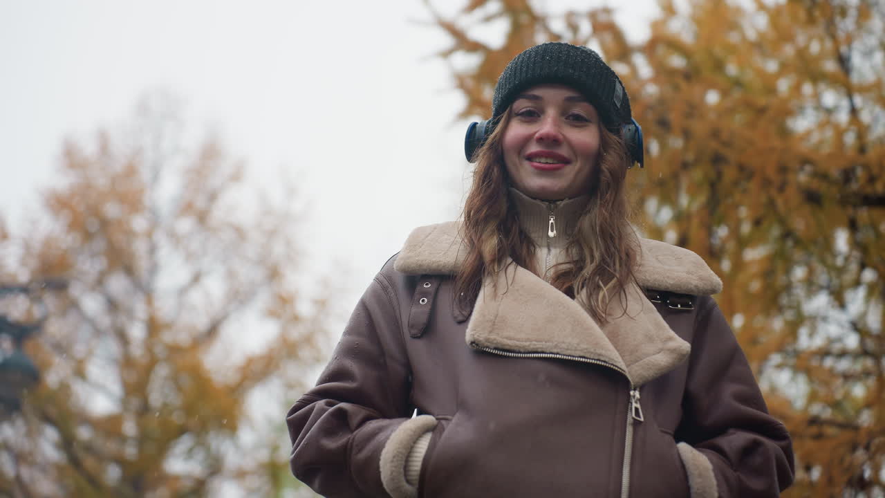 Smiling lady wearing black knit cap and brown shearling jacket with hands in pockets outdoors during cool autumn day, enjoying nature and fresh air