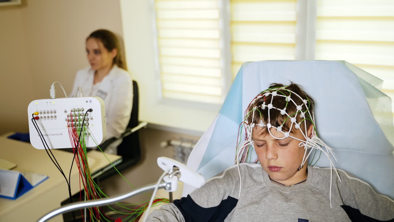 Teenage boy undergoing EEG examination in the hospital. Boy's head connected to lots of sensors. Laboratory medic at backdrop.