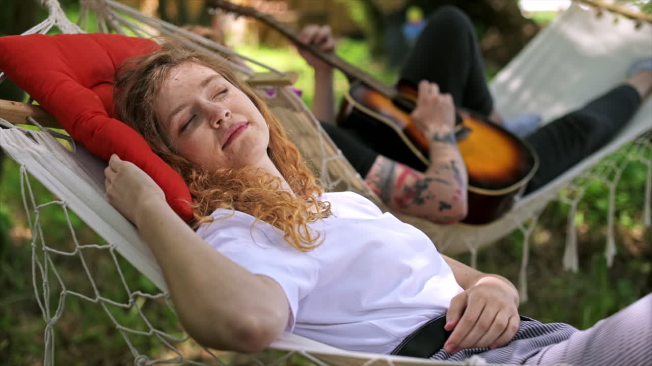 Couple lying on hammocks. Young man is playing guitar while woman is lying with closed eyes and smile. Greenery around. Glamping. Slow motion