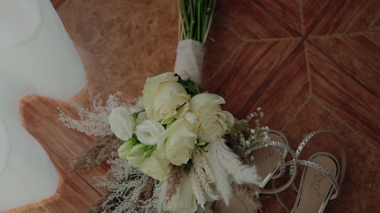 Elegant bridal bouquet with white roses and pampas grass resting beside sparkly sandals on a wooden floor