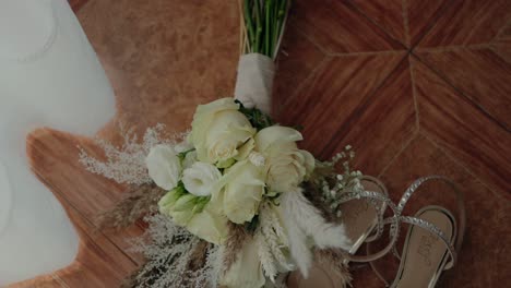 elegant bridal bouquet with white roses and pampas grass resting beside sparkly sandals on a wooden floor