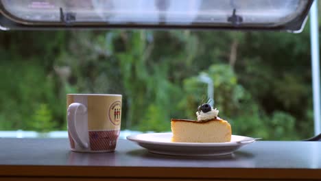 close-up steaming hot coffee or tea cup with cake on table with view on green forest. coffee cup with smoke in camper van with nature view. living van life on wheels