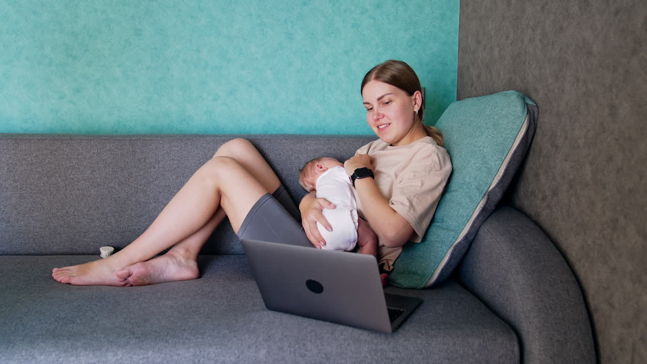 New-made mom with baby in hands sits on sofa. Caucasian woman working remote at home combining work and childcare.