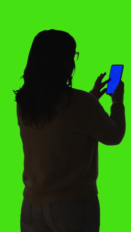 vertical video rear view silhouetted shot of woman holding blue screen mobile phone standing against green screen 1