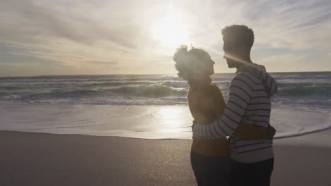 pareja romántica hispana de pie y abrazándose en la playa al atardecer