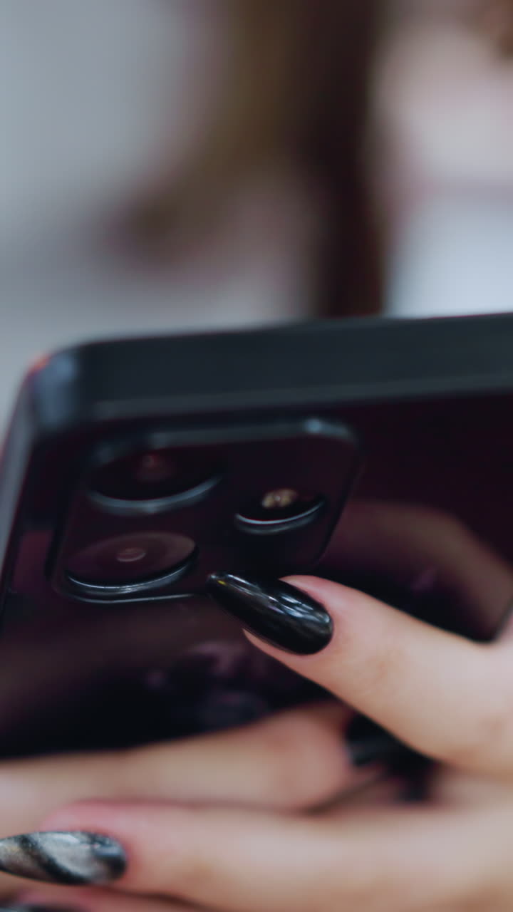 primer plano de un teléfono negro con uñas pintadas de mujer escribiendo un mensaje en el teléfono, la mano descansando en una mesa de madera marrón, rodeada por un fondo borroso suave