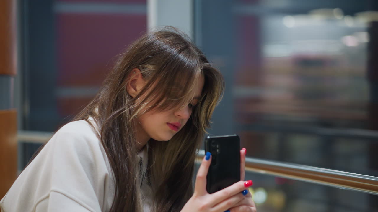 Young lady holding smartphone focused on taking photo of her classmate indoors near glass window while blinking city lights reflect across surface in modern shopping mall during evening