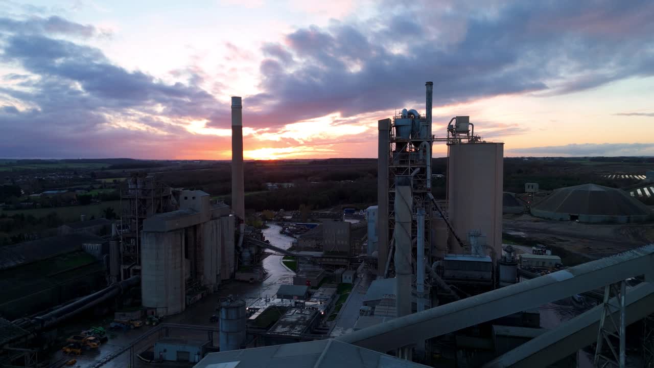 Volo cinematografico su un complesso industriale pesante con forni e infrastrutture di stoccaggio sotto la calda luce del tramonto.
