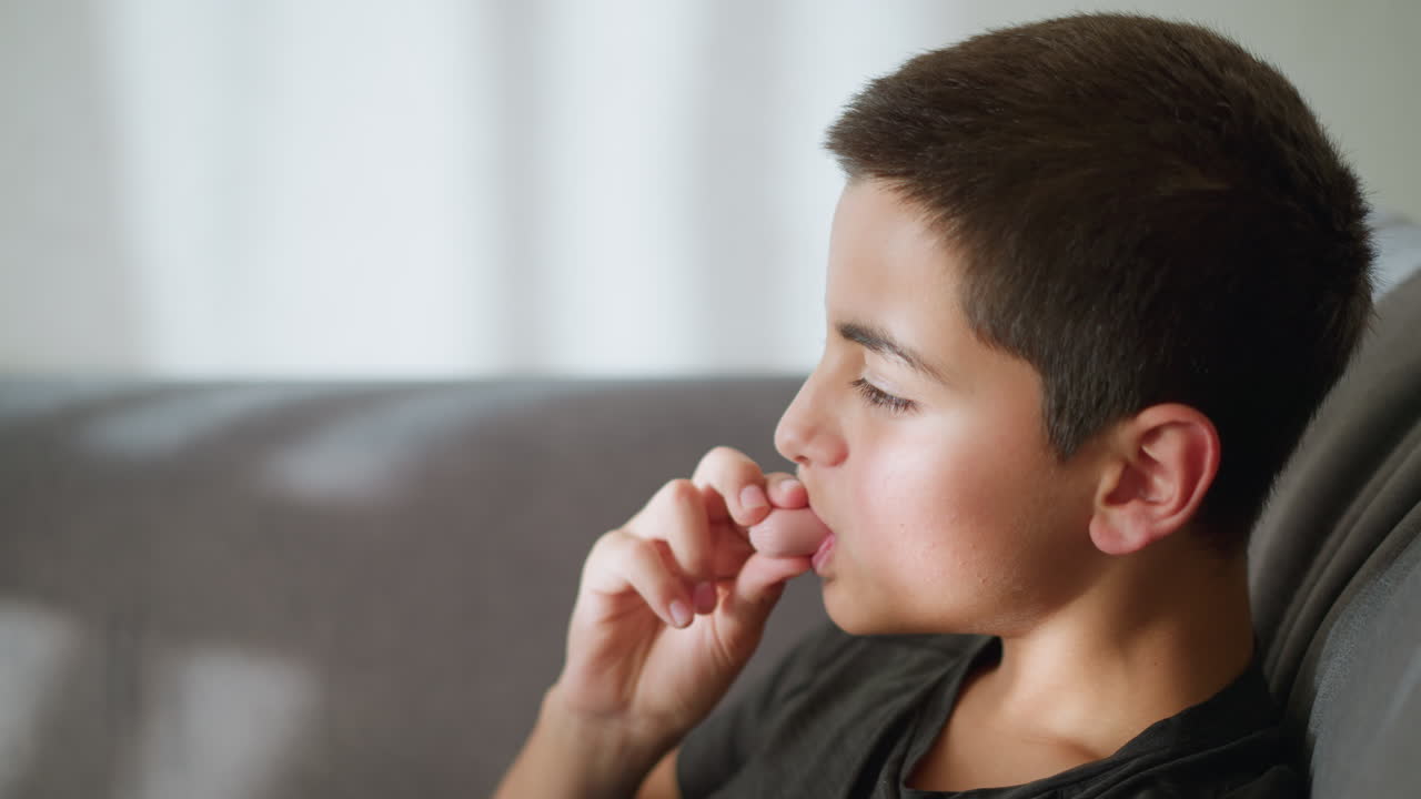 Close-up of young boy eating ice cream while sitting on couch, with light reflecting on his face as he enjoys the treat, the boy looks content and relaxed