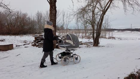 female mother slowly walk with baby carriage on snowy countryside road
