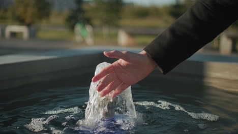 close up of man reaching out to touch flowing spring water from small fountain, with sunlight reflections on water surface and blurred outdoor setting including greenery and distant park bench