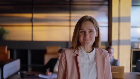 Portrait-of-a-confident-middle-aged-blonde-businesswoman-in-a-pink-business-suit-near-her-colleagues-at-her-desk-in-a-sunny-office