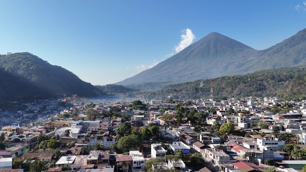 En fantastisk droneflyvning over San Lucas, Guatemala, viser tropiske kvarterer, lyse skyer, palmetræer og en dramatisk vulkanbjergkæde, der rejser sig over frodigt skovklædt terræn