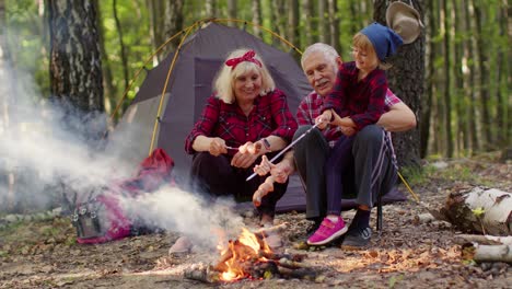 Senior-grandmother-grandfather-with-granddaughter-cooking-frying-sausages-over-campfire-in-wood