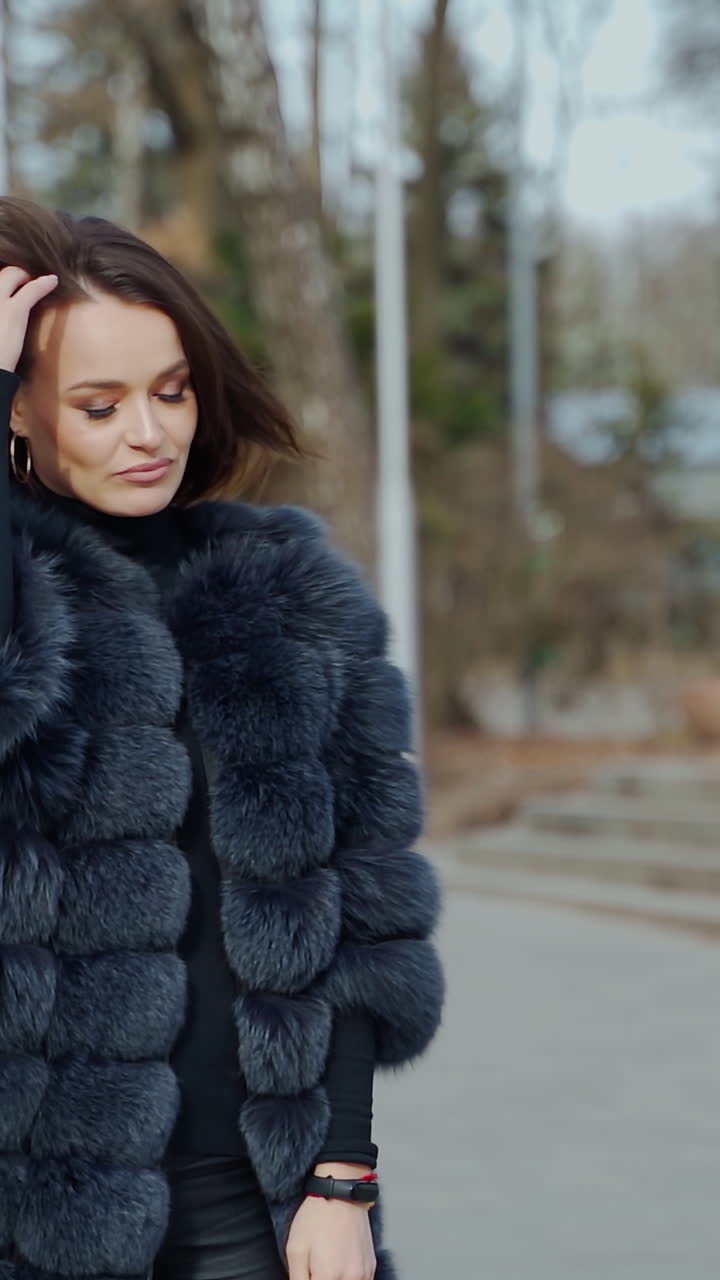 Street portrait of a fashionable woman. Young beautiful model wearing stylish black fur coat walking in the park. Model posing on camera. Street fashion concept. Vertical video