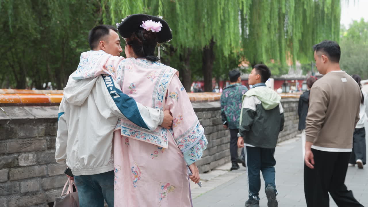 Coppia asiatica in hanfu che si abbraccia camminando lungo un canale di pietra fiancheggiato da salici, veste rosa pastello con fermaglio per capelli ornato, partner in giacca casual con un braccio intorno, palo della lanterna e ringhiera del parco, turisti di passaggio.