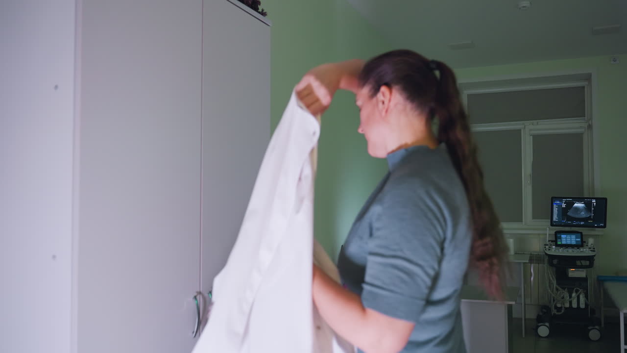 Female practitioner takes out uniform from cabinet in modern clinic, prepares for work by gently closing cabinet and putting on professional medical clothing beside desk and medical monitor equipment