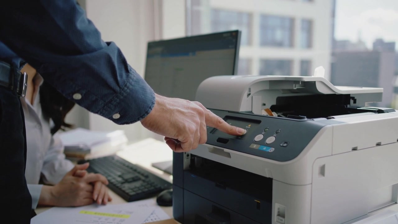Reaching man pressing printer control panel at office desk to copy papers, woman in blouse watching