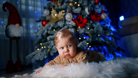 blue-eyed child with blond hair lies on the floor under the christmas tree. adorable baby crawls by the floor.