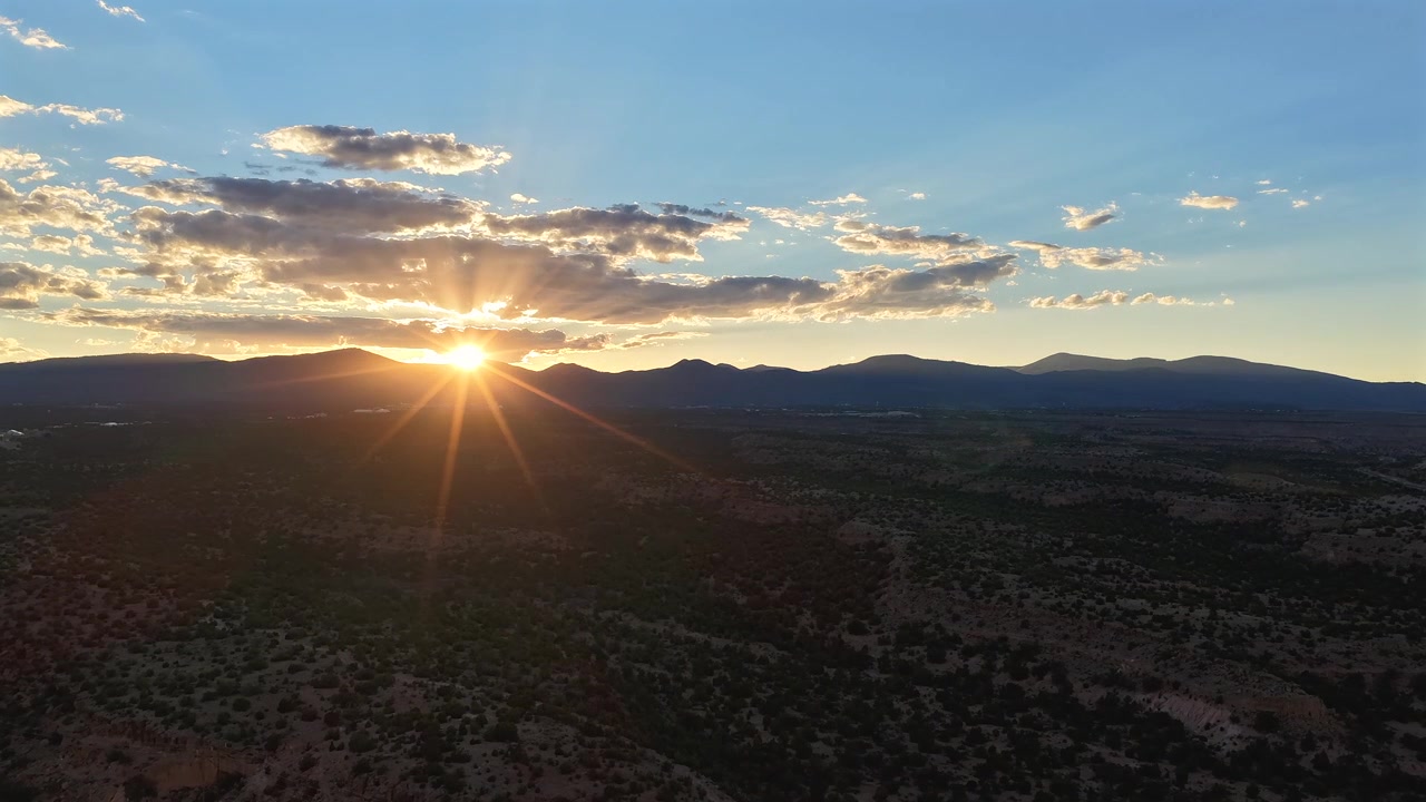 4K drone timelapse of a shifting southwest climate and cloud formations. A bright sunburst momentarily hides behind a dynamic weather system before setting behind a mountain horizon and vast mesas.