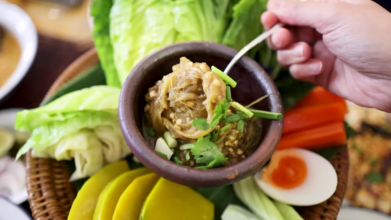 Hand uses spoon to scoop roasted green chili dip from clay bowl, surrounded by fresh vegetables and egg, in bright indoor Thai dining setting