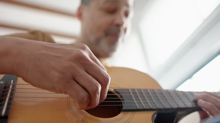 Inquadratura dal basso di un uomo anziano birazziale seduto che suona la chitarra a casa, messa a fuoco selettiva, slow motion