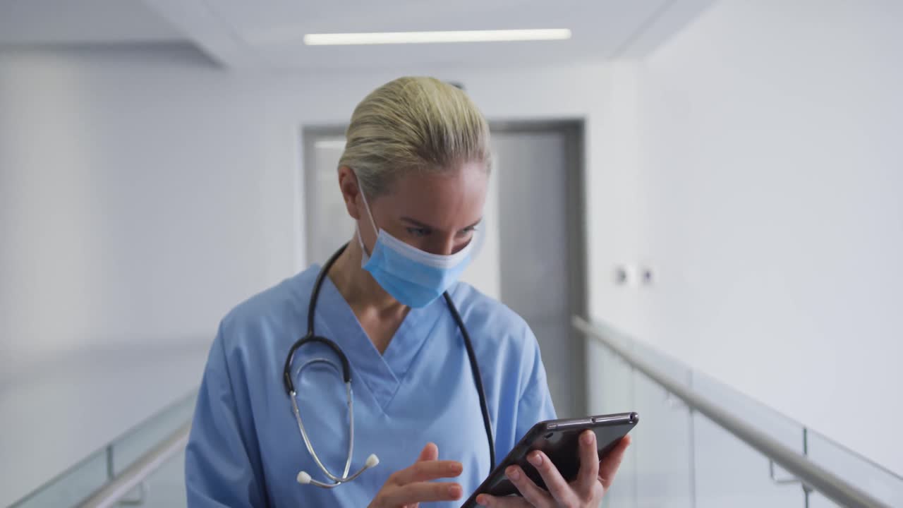 Doctora caucásica con mascarilla usando una tablet, mirando a cámara en un pasillo de hospital.