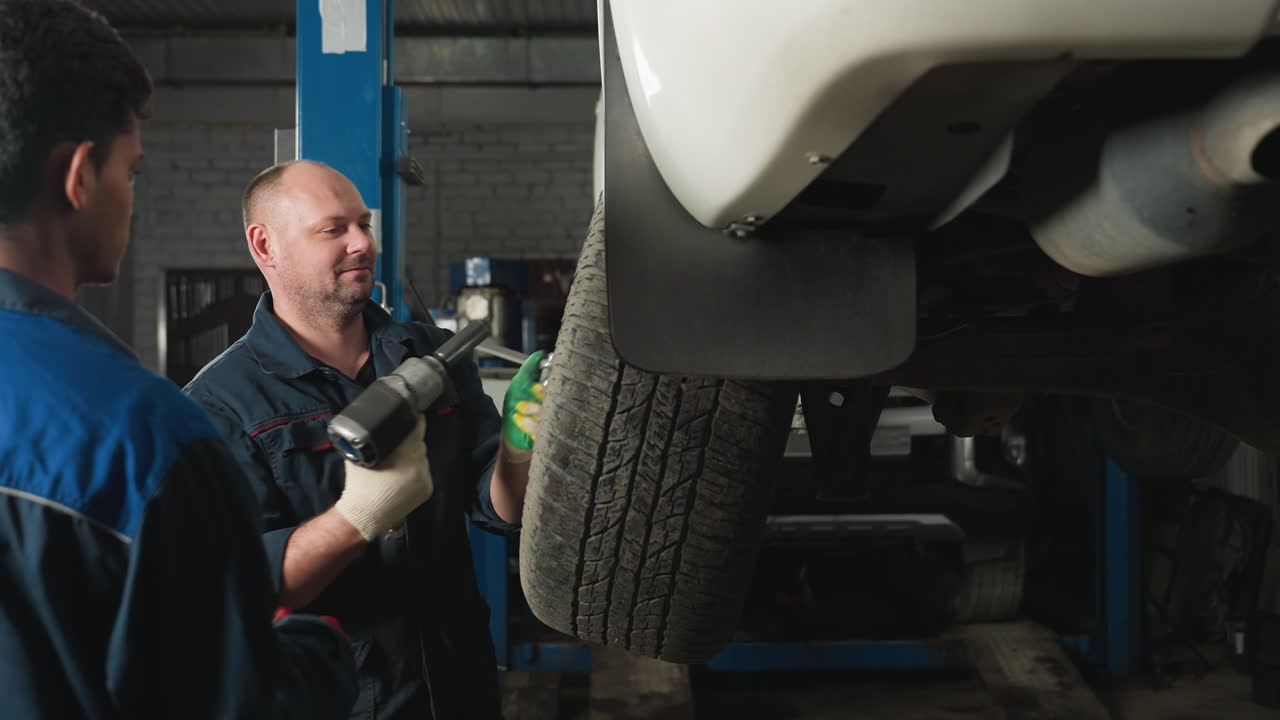 mecánico trabaja bajo el coche usando una llave mientras un colega sostiene la luz para ayuda, dos técnicos de laboratorio en el fondo, uno se vuelve para observar el trabajo que se hace en el taller mecánico