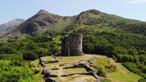 Aerial-shot-of-castle-ruins-in-Wales,-shot-on-a-sunny-day-with-the-beautiful-countryside-in-the-background-including-a-lake,-rolling-hills,-and-trees