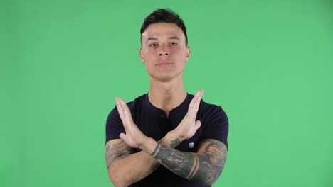 portrait of a handsome young guy looking at camera is strictly gesturing with hands crossed making x shape meaning denial saying no. green screen