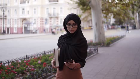 Eine-Junge-Muslimische-Frau-In-Einem-Traditionellen-Kopftuch,-Die-Mit-Freunden-Auf-Einem-Smartphone-Auf-Der-Straße-Chattet,-Während-Sie-Ihre-College-Bücher-Trägt