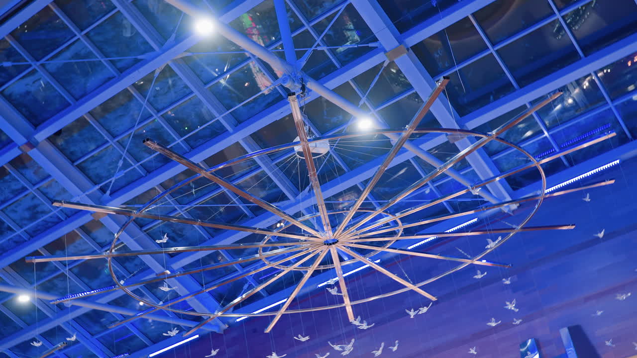 Rotational upward view of modern mall ceiling showing circular installation with spokes and decorative hanging birds under glass roof illuminated with blue lights