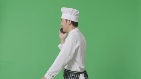 vista lateral del chef asiático hablando en el teléfono inteligente mientras camina en el estudio de fondo de la pantalla verde